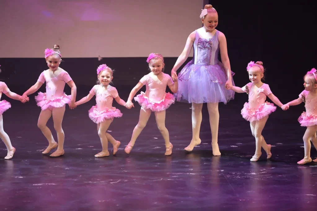 Ballet teacher in purple tutu leading tiny dancers in pink tutus holding hands on stage