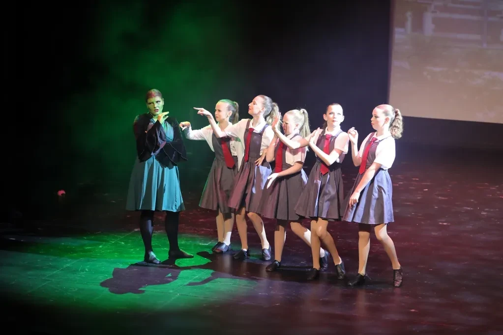 Wicked school scene with green witch and students in grey uniforms under green lighting