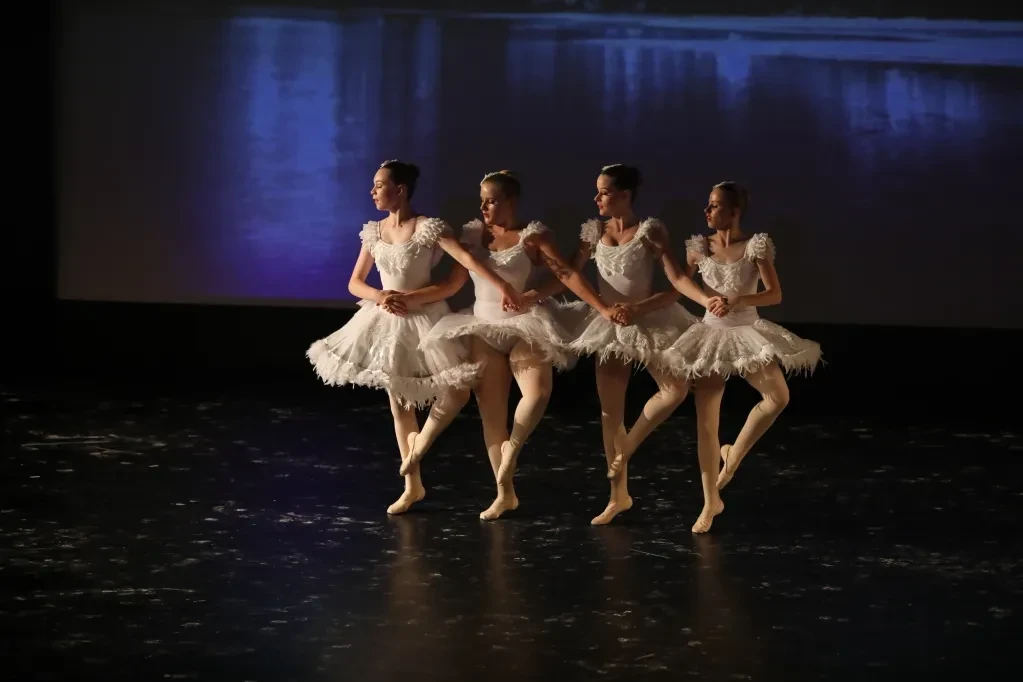 Four ballet dancers in white feathered tutus performing Swan Lake with lake backdrop