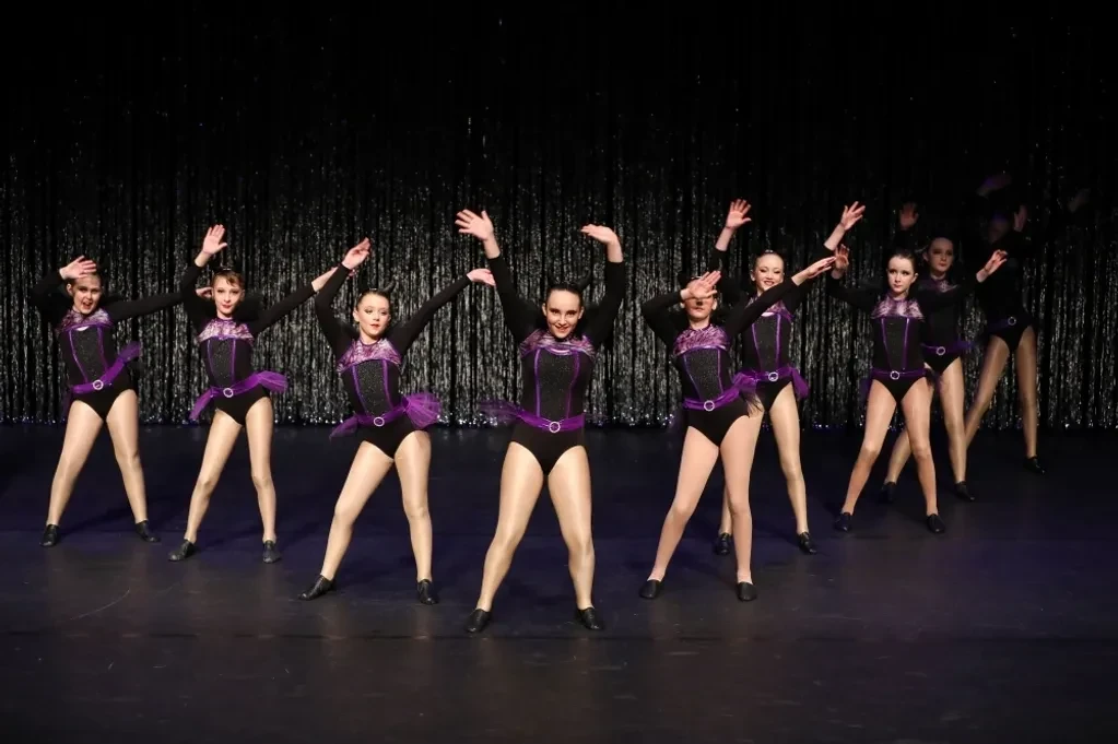 Eight dancers in black and purple sequin jazz costumes performing in a line against silver curtain