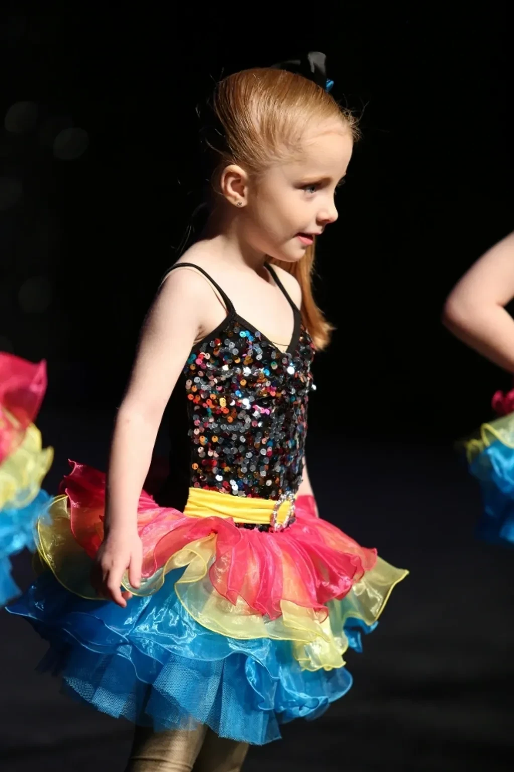 Young girl in black sequin top and rainbow ruffled tutu performing solo on stage