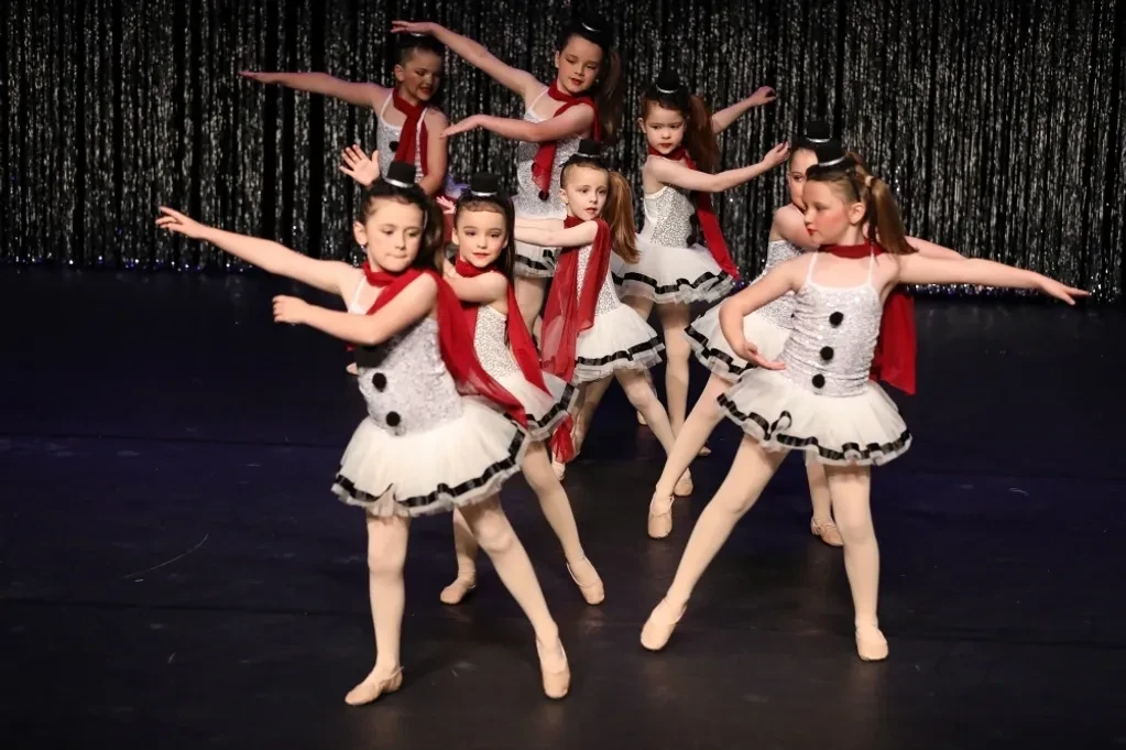 Group of children in white snowman costumes with red scarves performing arms-wide formation