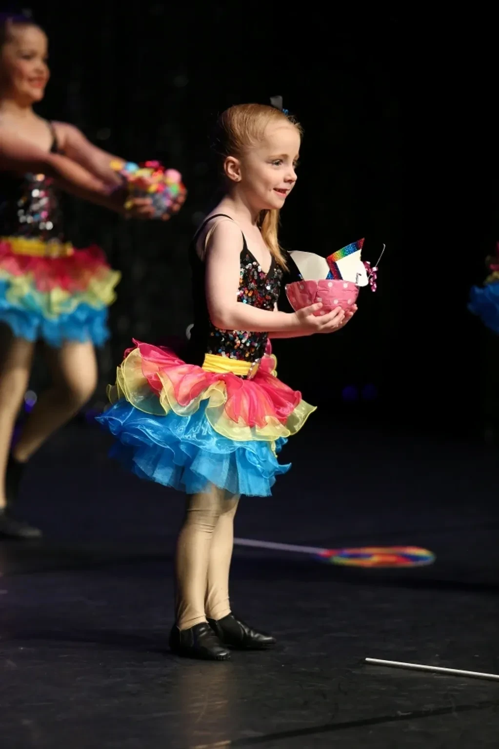 Young girl in rainbow tutu holding a pink cupcake prop on stage