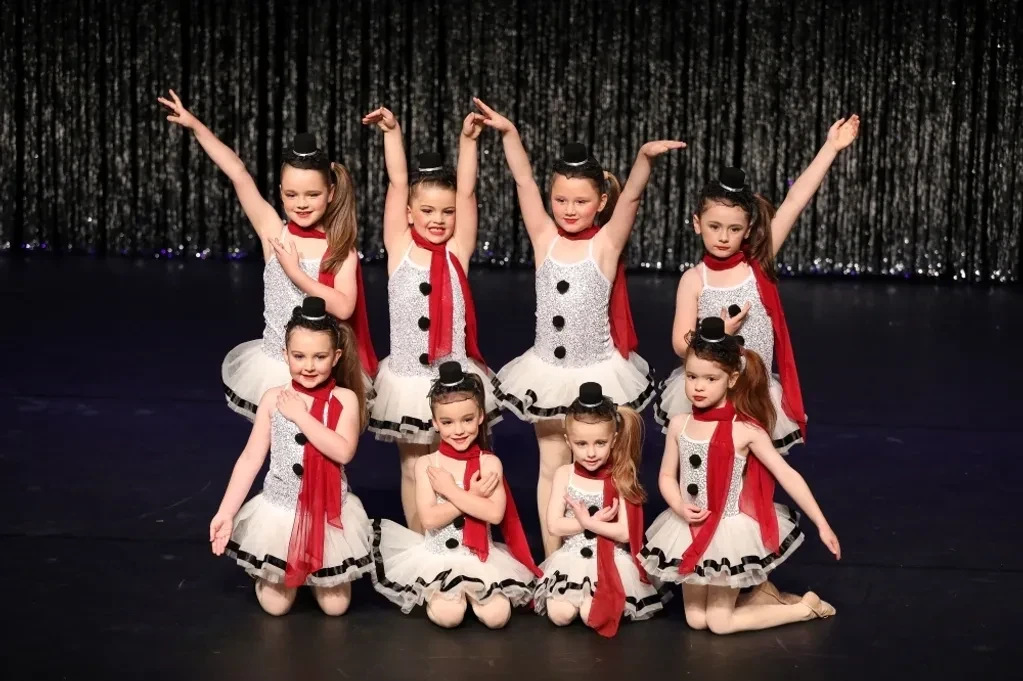 Children in white sequin snowman costumes with red scarves and top hats performing on stage