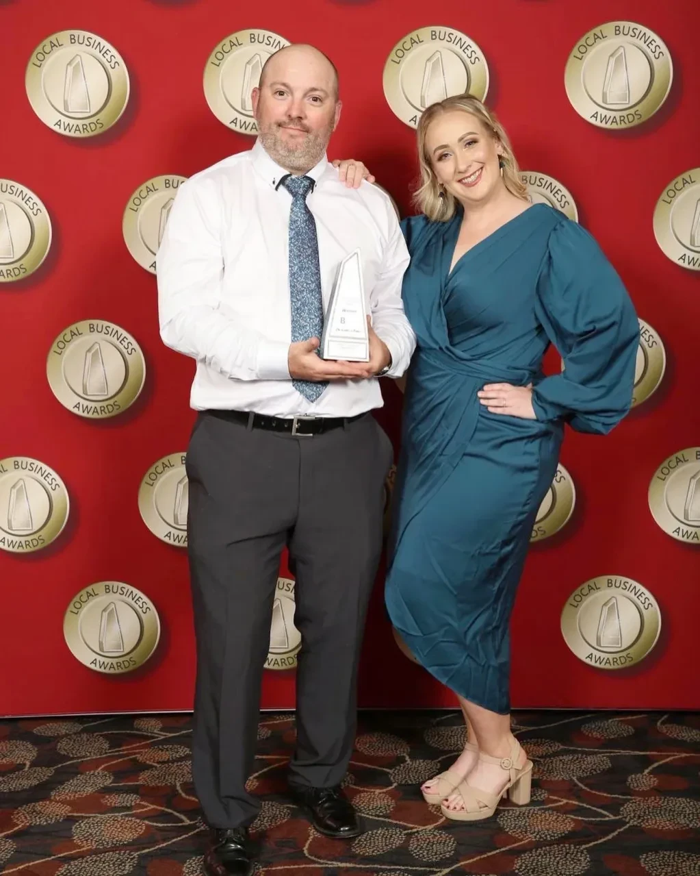 Man and woman holding Local Business Awards trophy in front of red awards backdrop
